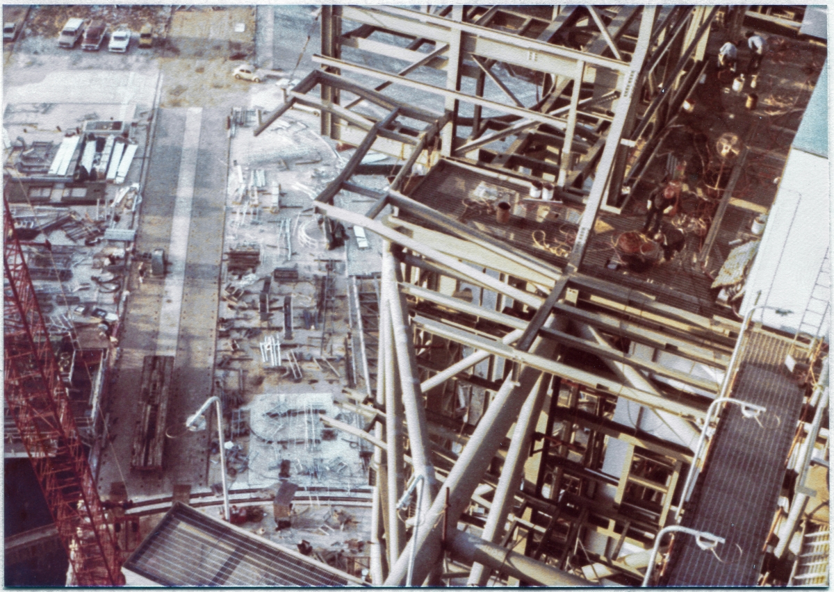 Image 008. Viewed from above on the Fixed Service Structure, the steel framework of the Rotating Service Structure at Space Shuttle Launch Complex 39-B, Kennedy Space Center, Florida, grows and takes shape as Union Ironworkers from Local 808, working for Wilhoit Steel Erectors, continue to work on it. In the distance, down on the ground, Wilhoit’s “shake-out yard” can be seen, straddling the western pair of tracks which are used by the Crawler, to transport the Mobile Launch Platform with the Shuttle on top of it to its launch position on the pad. Photo by James MacLaren.